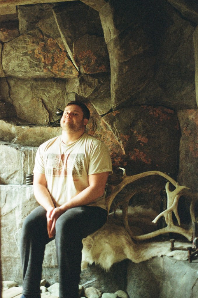A dashing man sits pulling a mischievous face looking at the camera next to reindeer antlers in a cavern. 