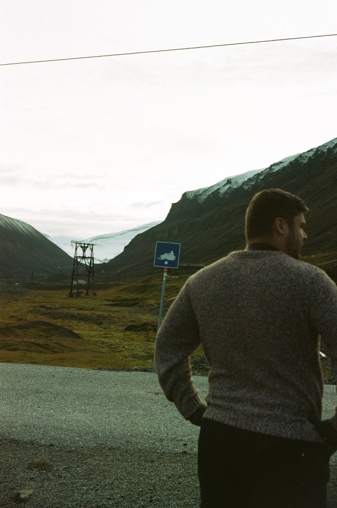 A man stands hands on his hips in the foreground with a signpost showing a snow mobile visible over his shoulder. 