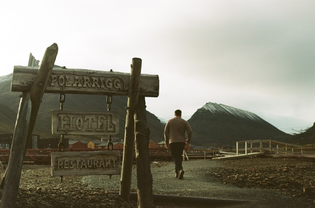 A wooden sign for the hotel shows 'Polarrigg. Hotel. Restaurant' A man is seen walking away. The town is seen in the background with a snow sapped mountain behind it. 