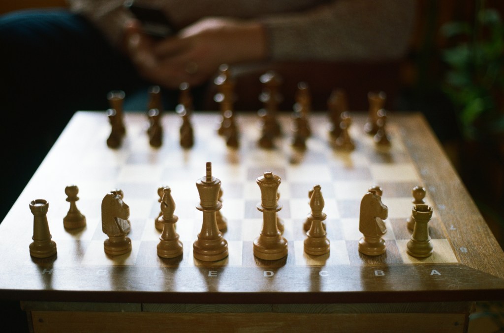 A chess board is set up ready to play a game, in the background the hands of the opponent are blurred. 