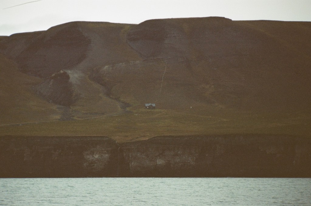 A mountain ending in a cliff that drops to the ocean with a house is seen. To he right of the house is small white cross. 