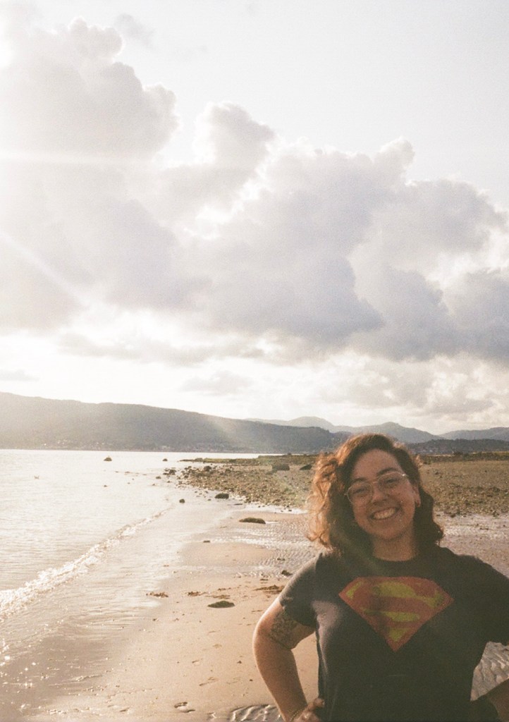 A 30 year old female stands with her hands on her hips smiling into the camera on a sunny beach. She is wearing a superman t-shirt and has smudges of paint on her face and shirt.