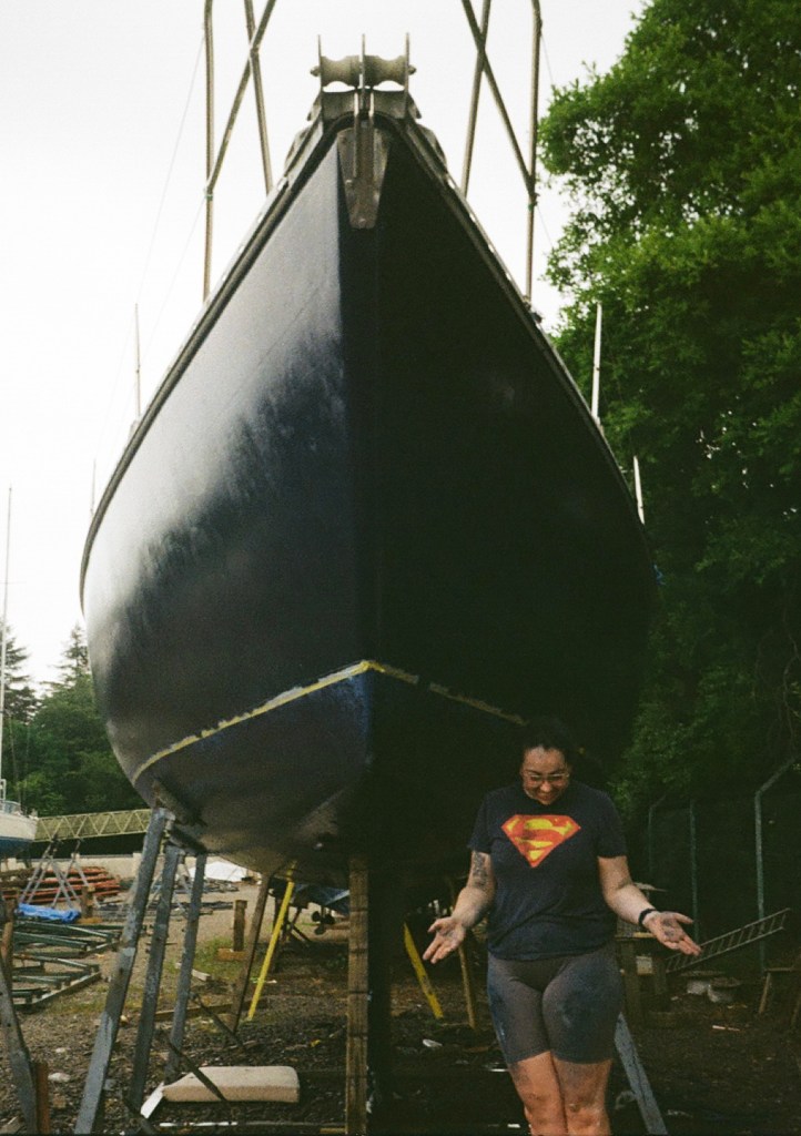 A woman stands in front of a painted boat looking down with her hands out palms up. She is smiling. Her clothes and skin are covered in paint. 
