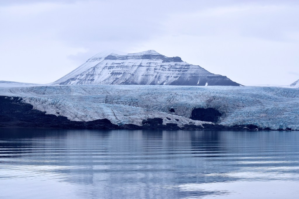 A glacier is seen reflected in rippling water. There is a large snow capped mountain in the distance.