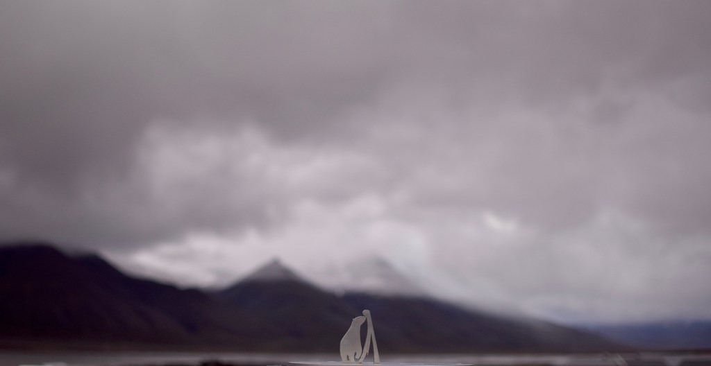 A small polar bear and mountain sculpture on a sun dial are in focus in the middle of the image. Snow capped mountains can be seen in the background. 