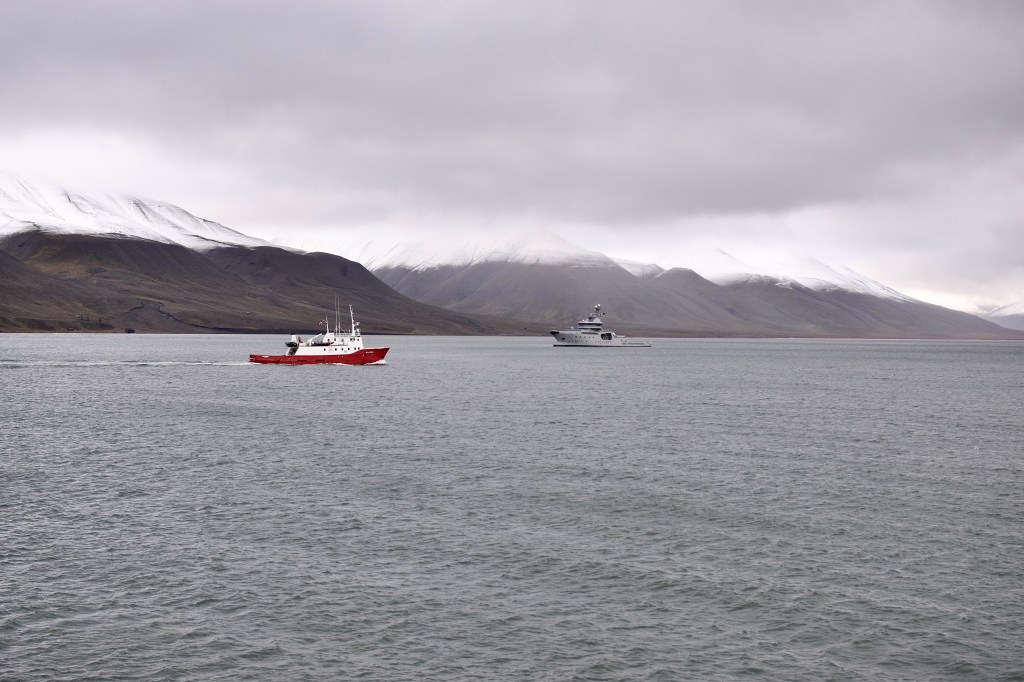 A body of water with 2 ships on it can be seen. In the background there are snow capped mountains. 