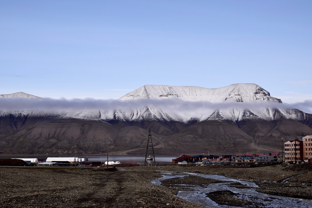 Scenic image showing snow capped mountains the other side of a fjord. The sky is blue. In the mid gorund is industrial looking buildings and a flats to the right of the image. 