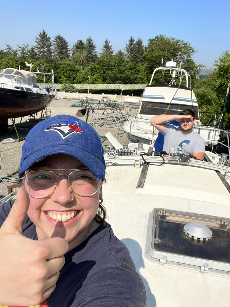 Sat atop a yacht on the hard a 30 year old female take a selfie with a thumbs up and a large grin, in the background a 30 year old male shades his face and looks towards the camera. He is smiling but appears bemused.