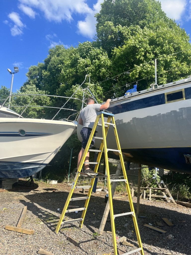 A man stands on a step ladder and is painting a boat. There is grey primer paint covering part of the hull. 