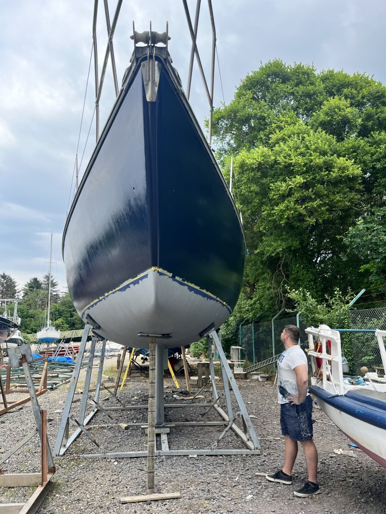 A man stands in front of a boat looking up at it with his hands on his hips,, His clothes are covered in paint. 