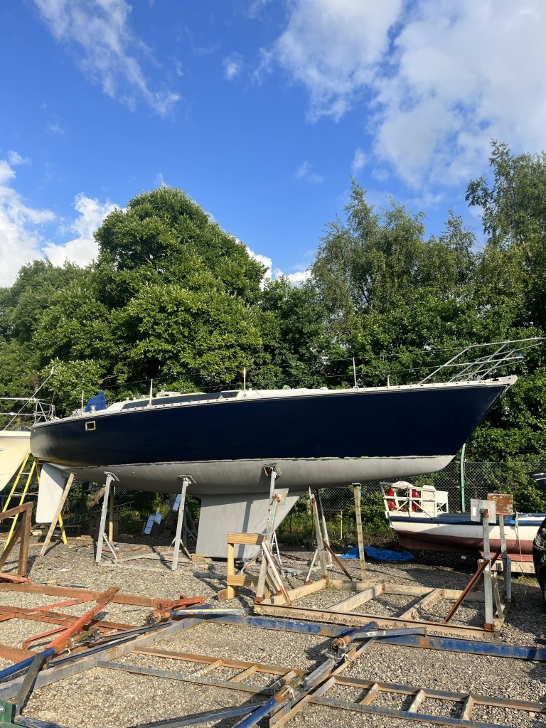 A full view of the side of a yacht in a cradle can be seen. The upper hull is a deep blue and has a shine. the lower part of the hull is a grey/ silver colour painted primer. There is a yellow line of masking tape splitting the two colours. 