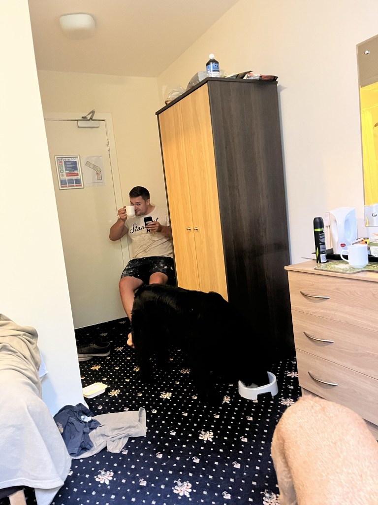 There is a room decorated possibly 20 years ago and mismatching furniture. Looking towards the door and in the corner there is a 30 year old male sitting gesturing cheers with a cup of tea. A black outline in front shows a flatcoated retriever eating his dinner from a bowl. The room is slightly messy.