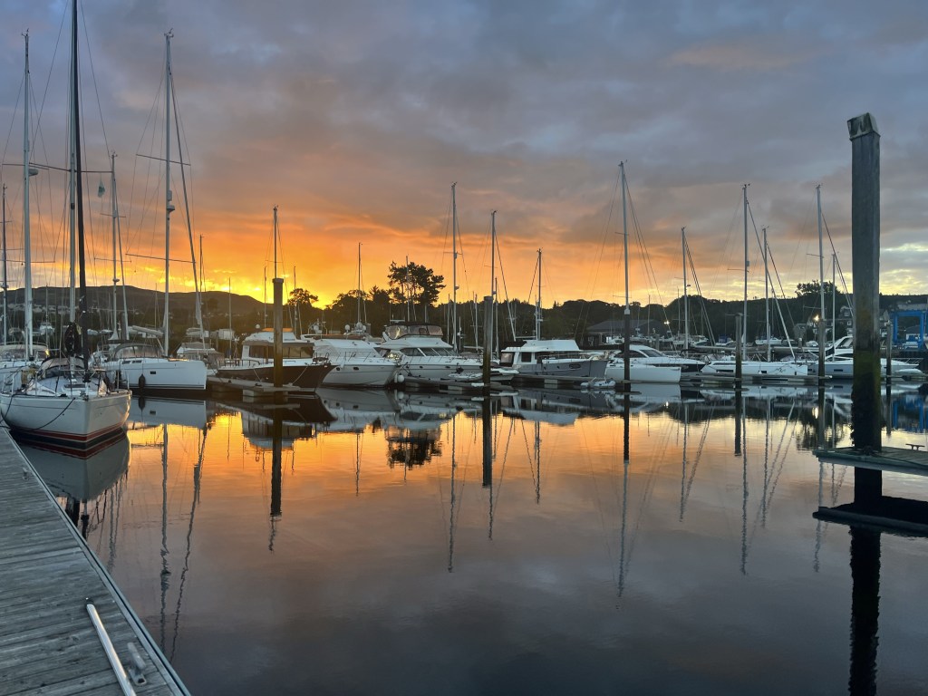 There is a view over the marina with yachts and part of a pontoon in the foreground. The water in the marina makes a perfect mirror reflecting the yellows and orange of the sunrise.