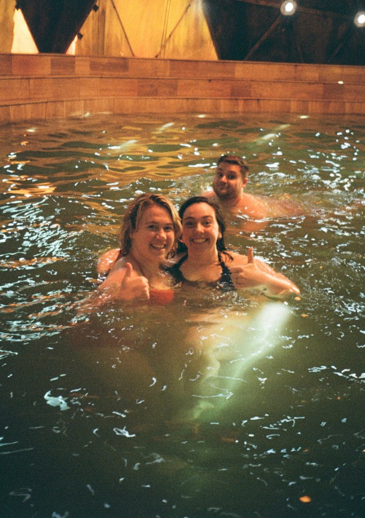 Two women give thumbs up whilst giving each other a hug in a pool, a man swims across in the background.