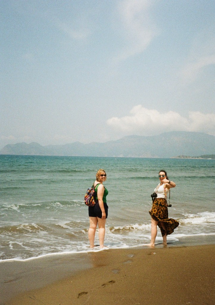 A blond woman and a brunette stand on a beach looking back towards the camera. The brunette has two cameras and is giving the thumbs up. The image has an old school film grain look to it.