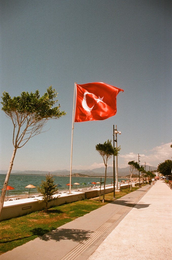 A turkish flag flies in the wind. The beach with deck chairs and umbrellas can be seen behind.