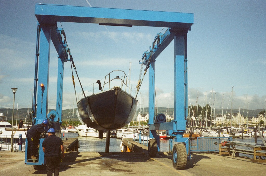 A boat is in a sling moving toward the edge of the marina water edge. 