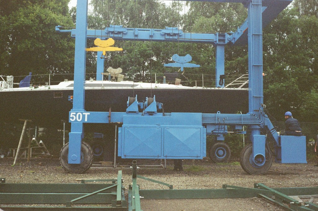A large blue winch system can be seen moving along the side of a boat that is sitting on its cradle on the dry ground. 