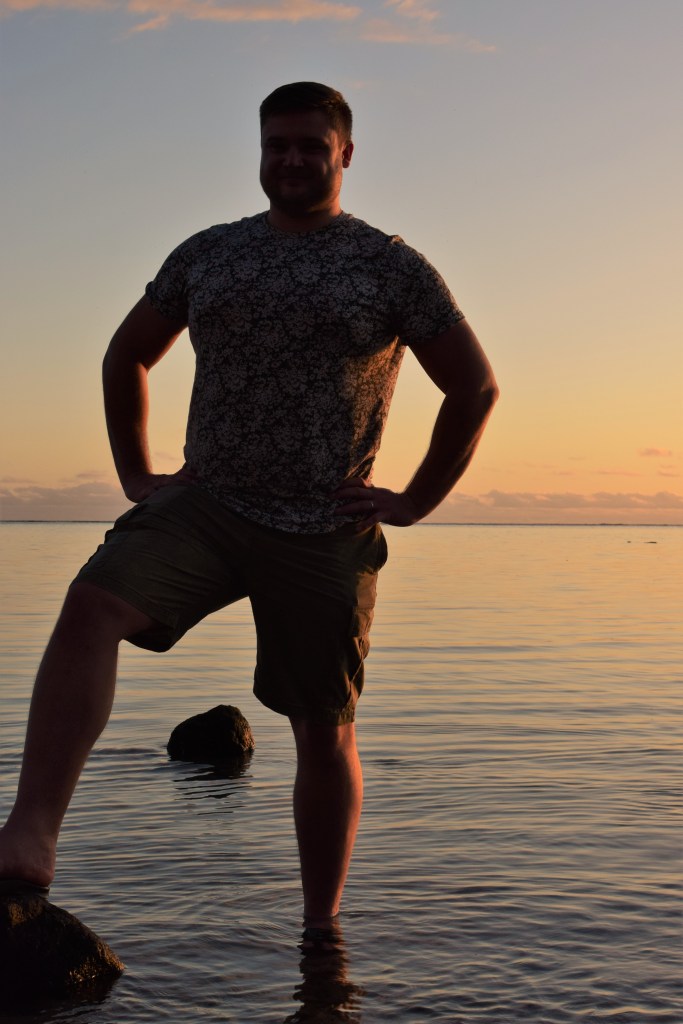 A man is standing in front of a sunset smiling into the camera. In the background between his legs is a rock in the water.