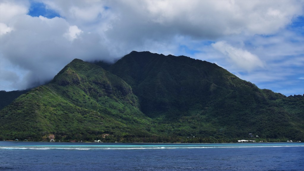 A lush green island with mountains with a ring of sand bank and corals.