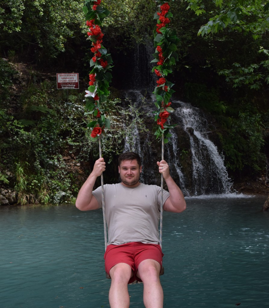 A man sits on a swing over water with a waterfall behind them.
