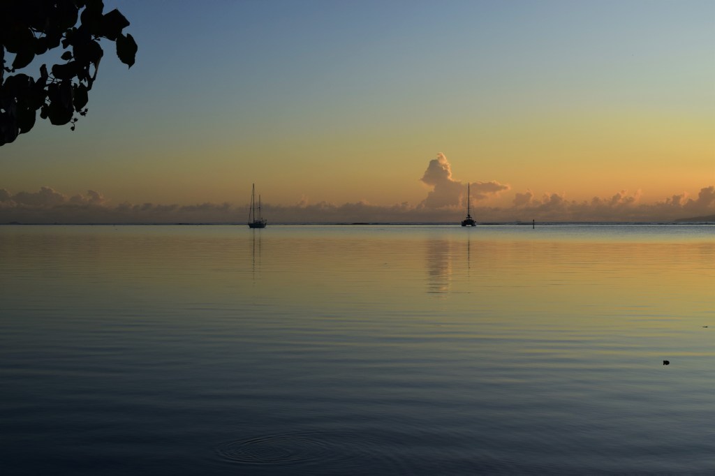 The pastel blue, green, yellow and pinks of a sunrise can be seen relelcted in clear water. Two yachts are also reflected in the water. 