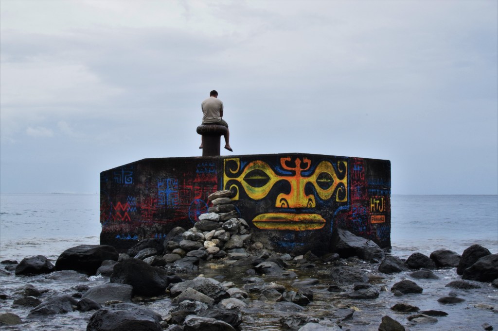 A man sits on a vibrantly graffitied pillar box in the sea.