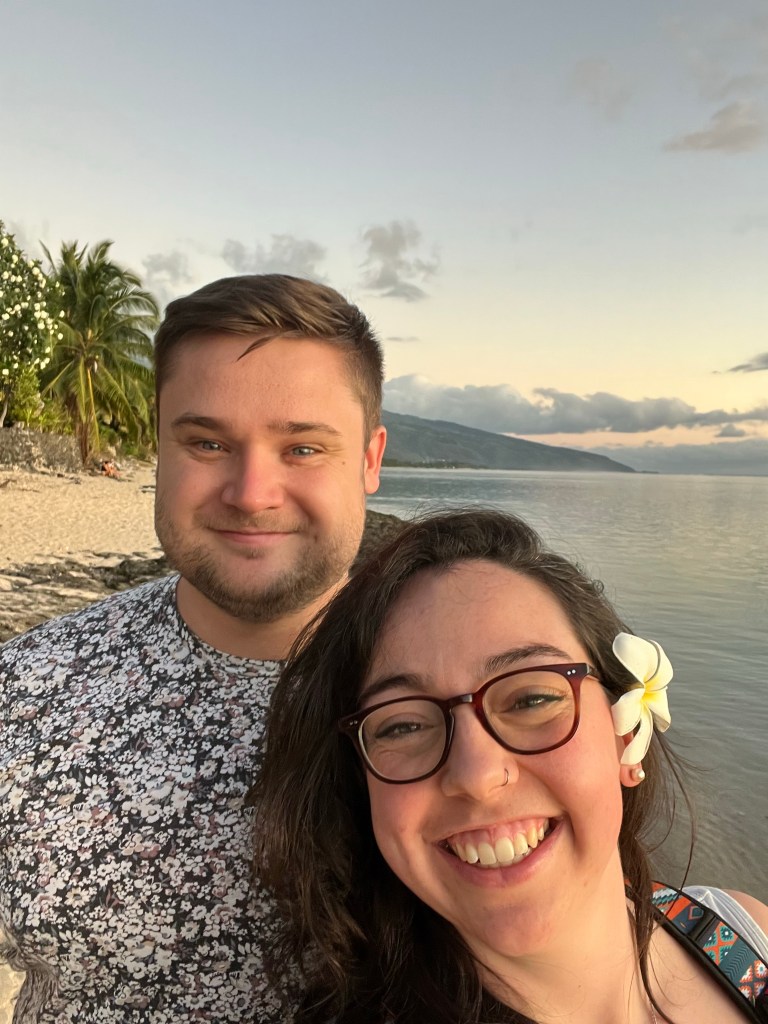 Brunette female and handsome man smile into the camera, in the background can be seen a beach, early sunset and the coast line. 