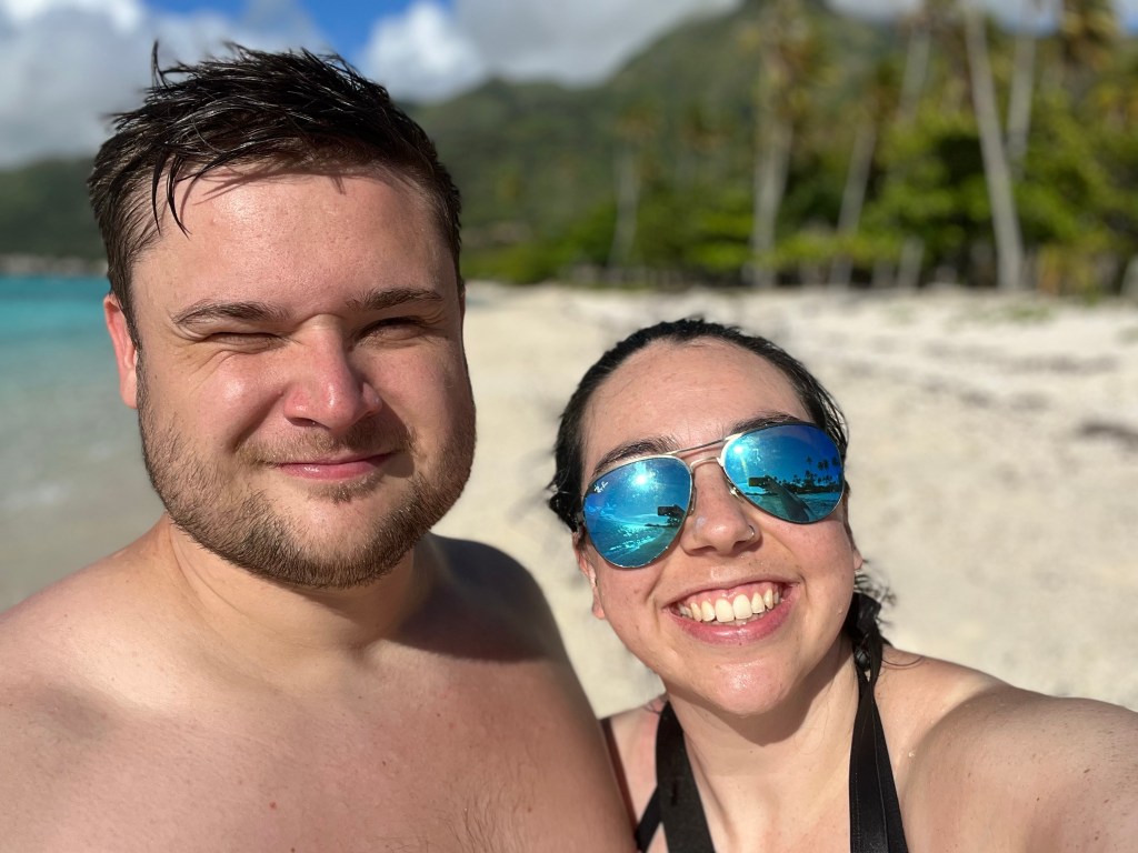 A woman in sunglasses smiles at the camera, she is next to a man squinting and smiling at the camera. In the background is a white same back and blue seas. 