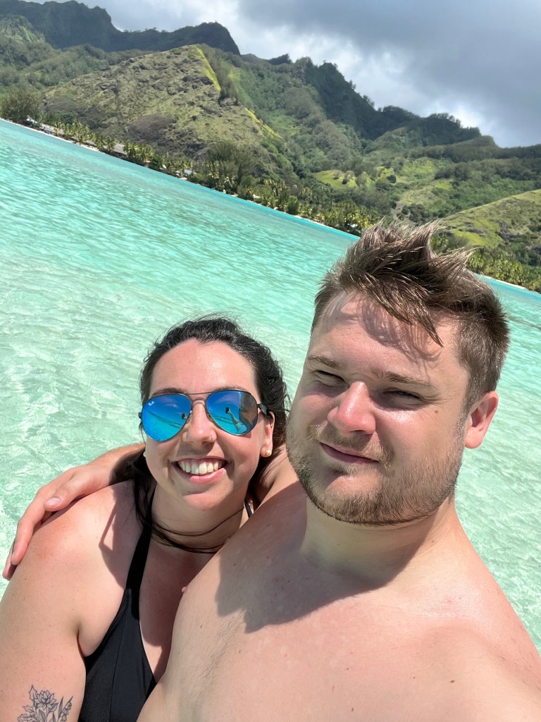 A man and woman take a selfie in swimwear in front of clear blue waters.