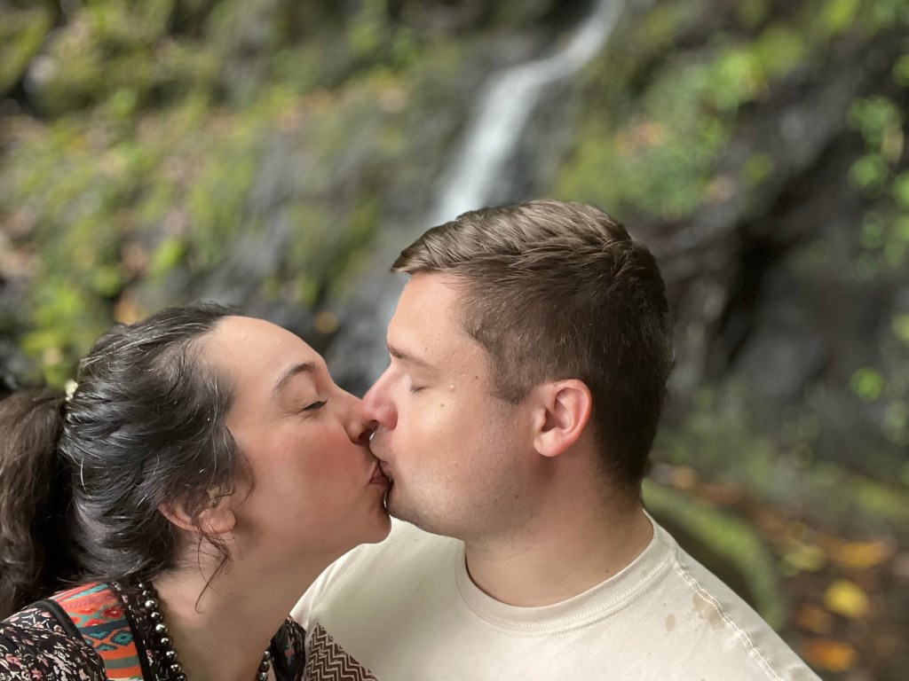 A man and woman kiss in front of a waterfall.