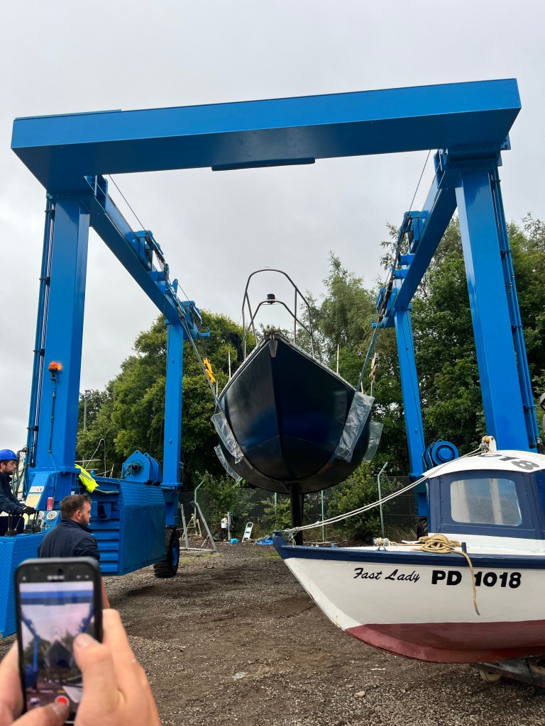 A boat in a sling is moving towards the camera view, to the left is a picture of a phone screen which shows the same picture being videoed. 