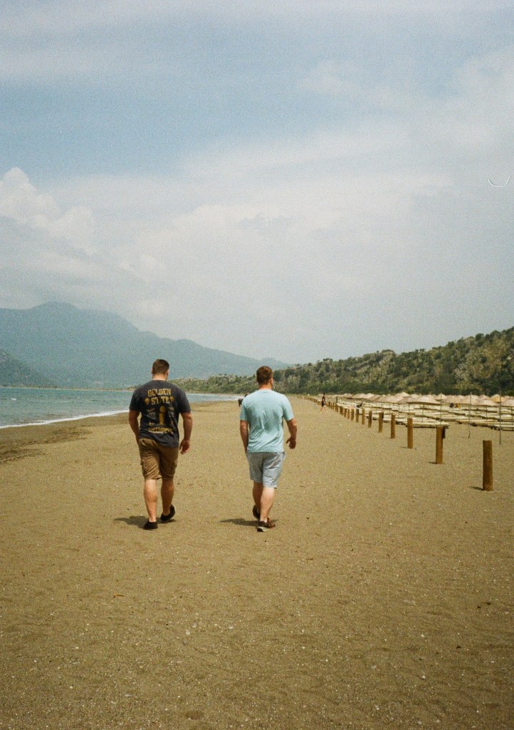 Two men walk away from the camera along a sandy beach.