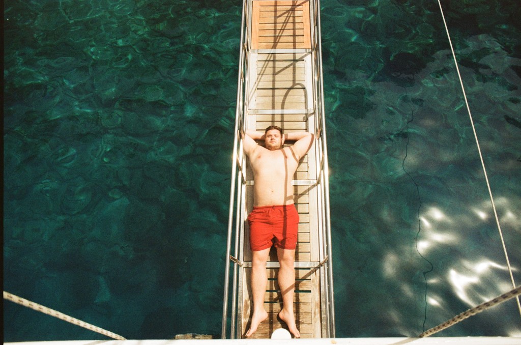 A man lays on a wooden walkway with his arms behind his head sunbathing.