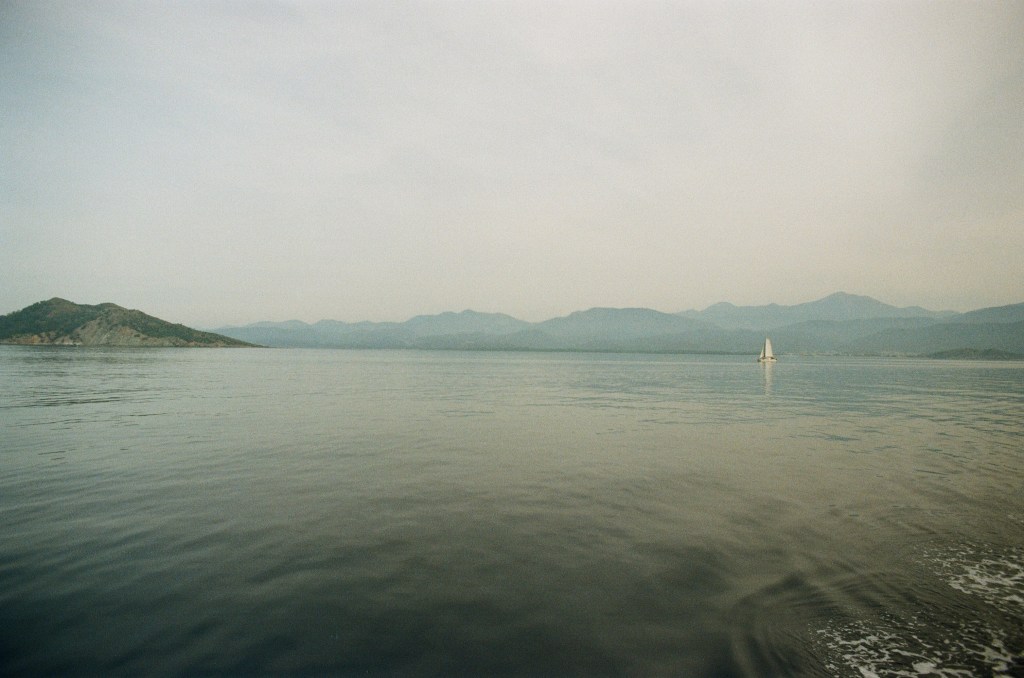 A still expanse of water with a sail boat can be seen in the distance, the film gives a cool tone to the image.