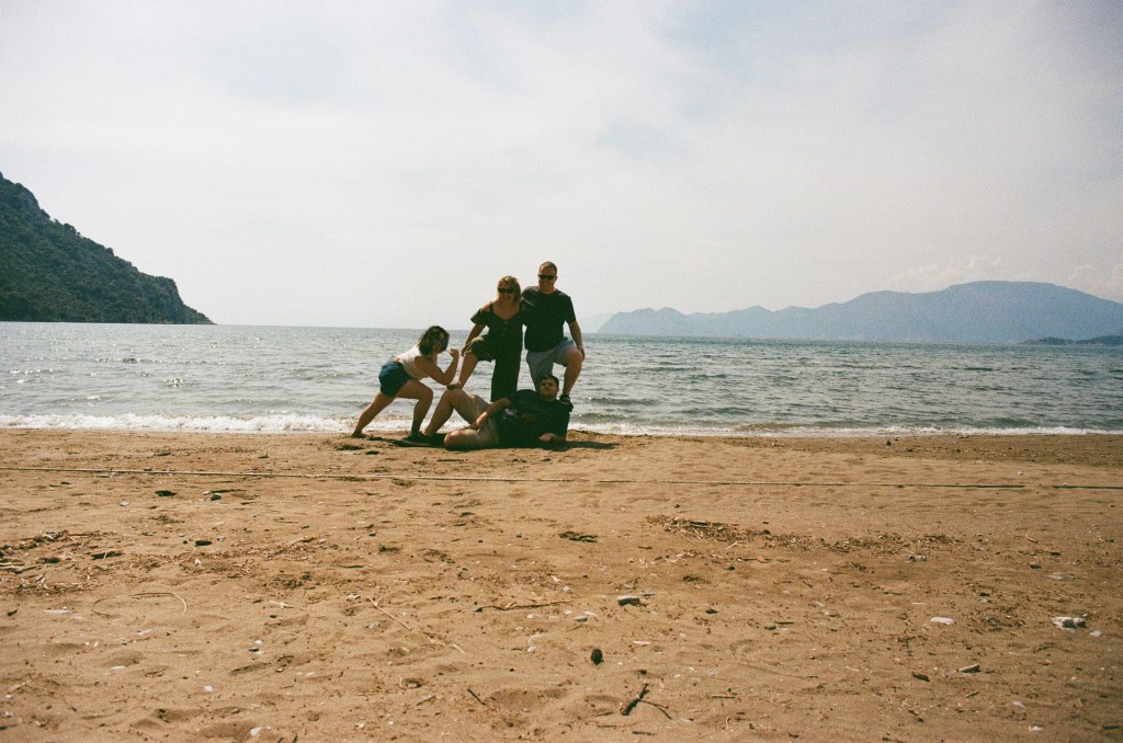 A group of 2 men and 2 women pose on the beach. One lays on the ground as 2 others put their feet on them and the last does a lunge. They're on a sandy beach.