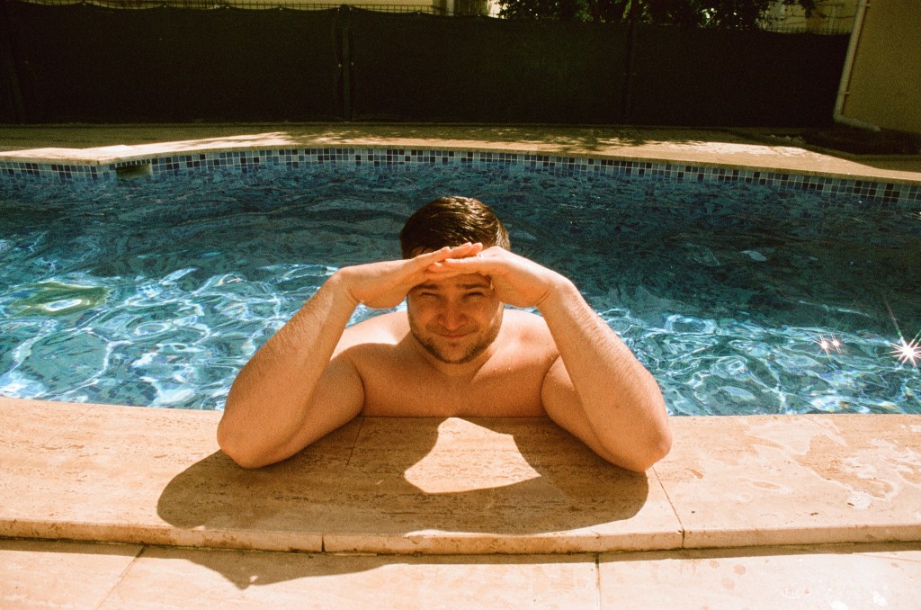 A man leans on the pool edge with his hands shading his eyes.