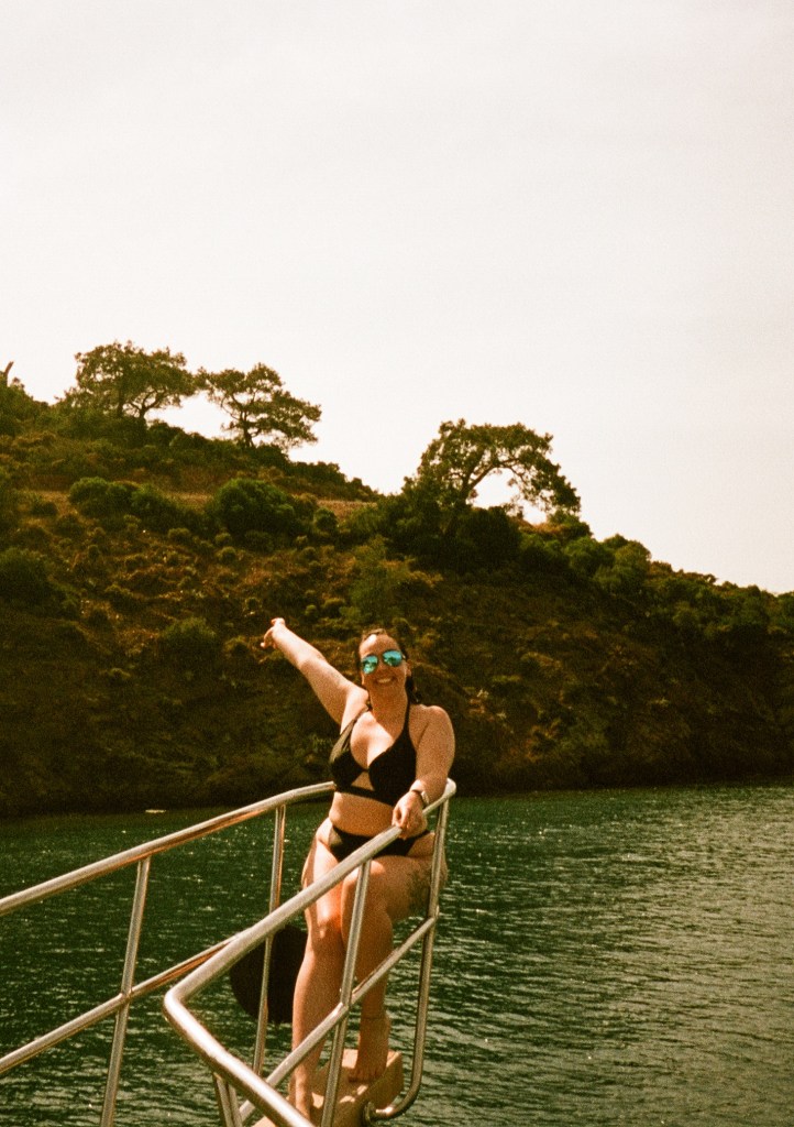 A woman sits at the front of a boat one arm outstretched.