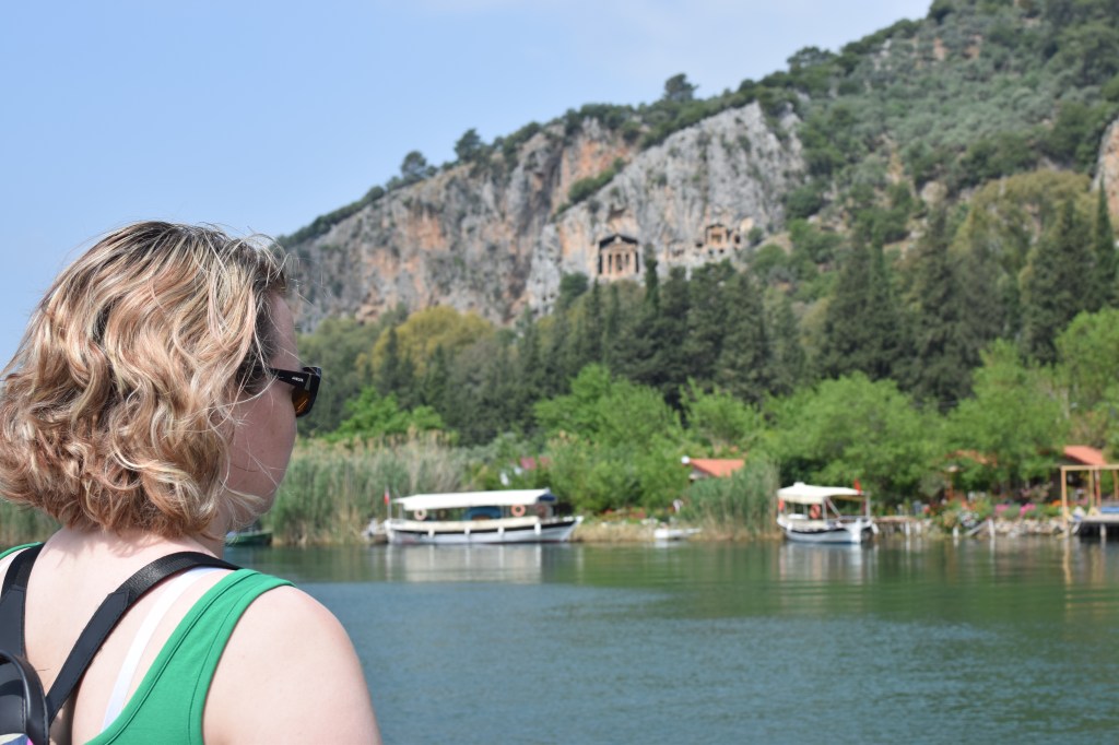A woman with short blond hair looks out across of body of water. In the background tomb fronts can be seen cut into a rock face and boats across the water.