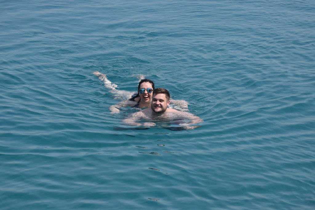 A brunette woman and man are swimming in water, the woman is partially on top of the man.