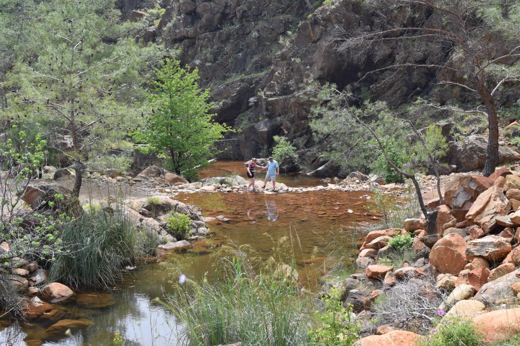 A blonde woman and blonde man walk across a gravel bath bordered by water.