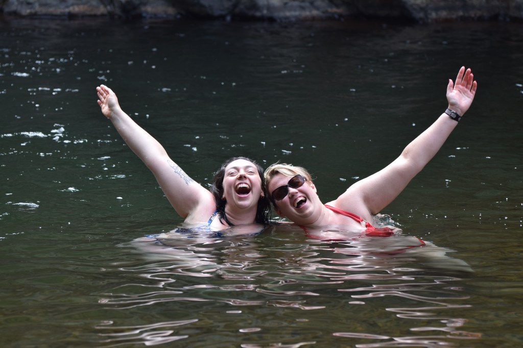 A blonde woman and a brunette woman in water have an arm outstretched each to the side. They are smiling.