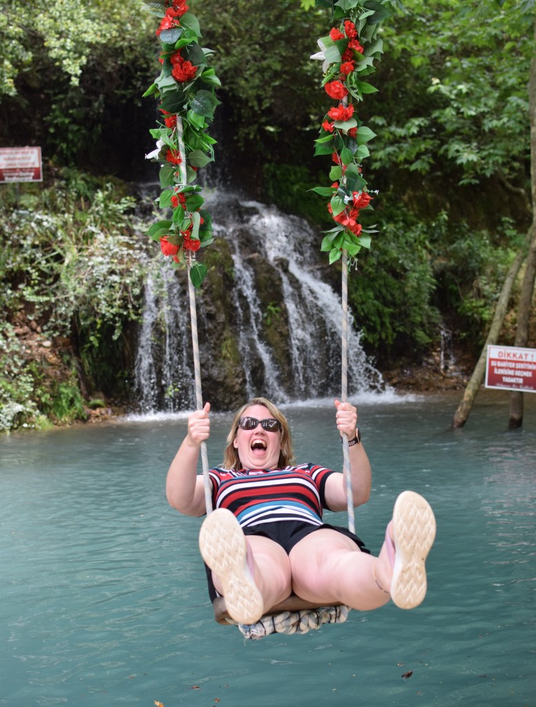 A blond woman is pictured swinging towards the camera over a river bed, there is a small waterfall behind her. She has a large open mouth grin.