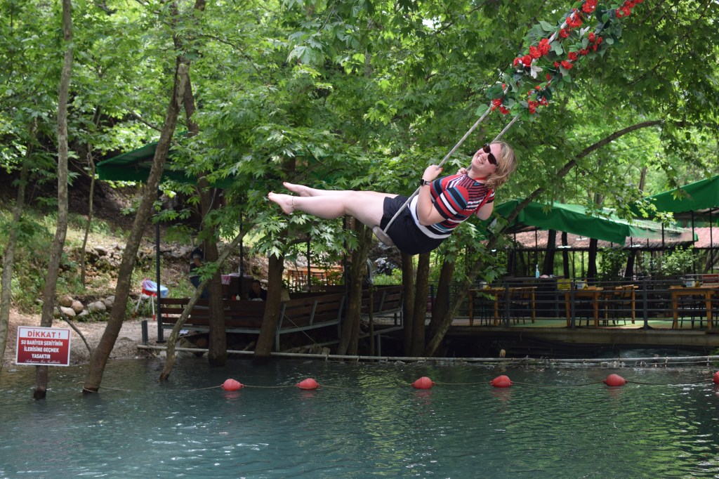 A blond woman is pictured on a swing from the side profile, she is a a long away above th river bed beneath her.