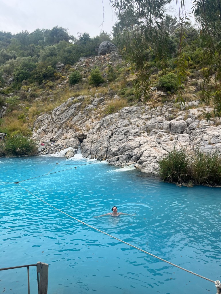 A woman is swimming in blue waters, behind there is a rocky edge to the lake.
