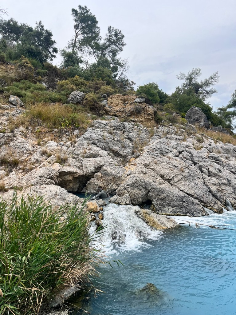Blue waters can be seen with a rock formation coming from the water to the left with a small waterfall. In the midst of the rocks a man is seen covered in mud.