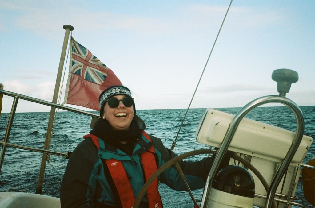 A woman in sunglasses, coat and life jacket sits at the back of a boat at the wheel. She is laughing. 