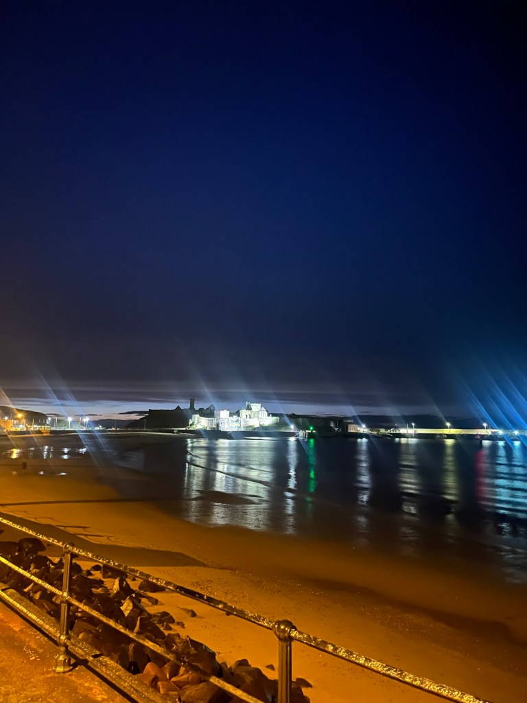 Shows a night time view of a castle and light reflected on water. This is in Peel. 