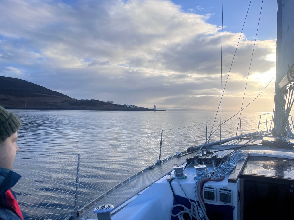 A man squints into the sun sitting to the side on a sailing yacht. The water is calm and there is an outcrop of land to the left with a lighthouse on the end.