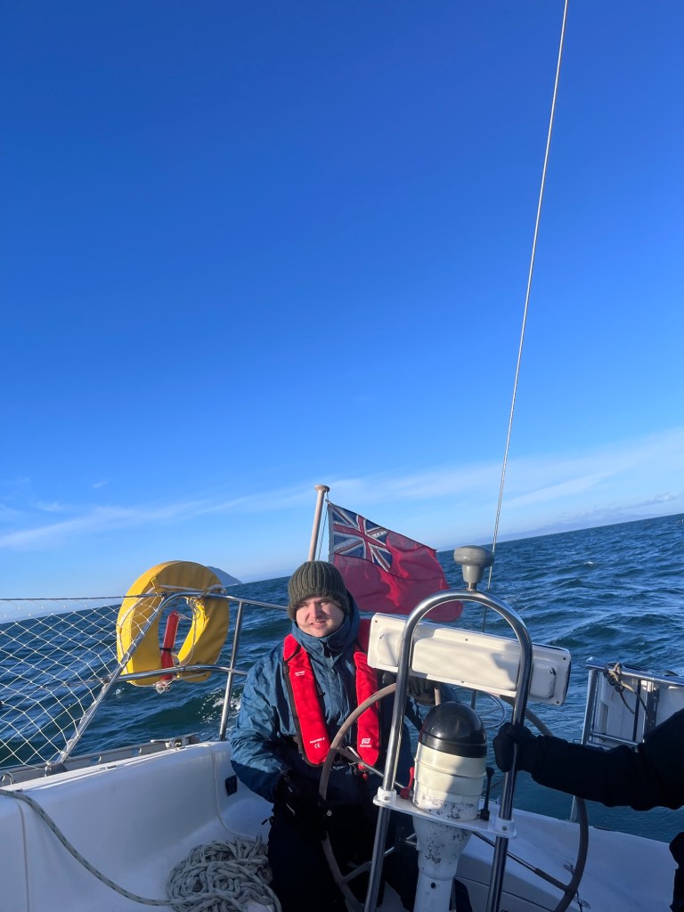 A man wearing a knitted hat, blue jacket and life jacket sits at the helm an outcrop of land can be seen in the distance. The yacht is obviously at an angle to the horizon behind him. 